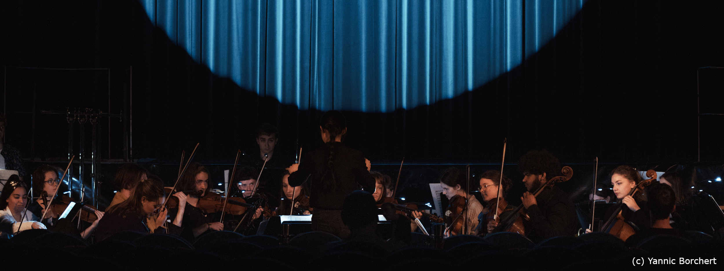 Orchester spielt im Vordergrund, im Hintergrund ist ein kreisförmiger Ausschnitt eines blauen Bühnenvorhangs