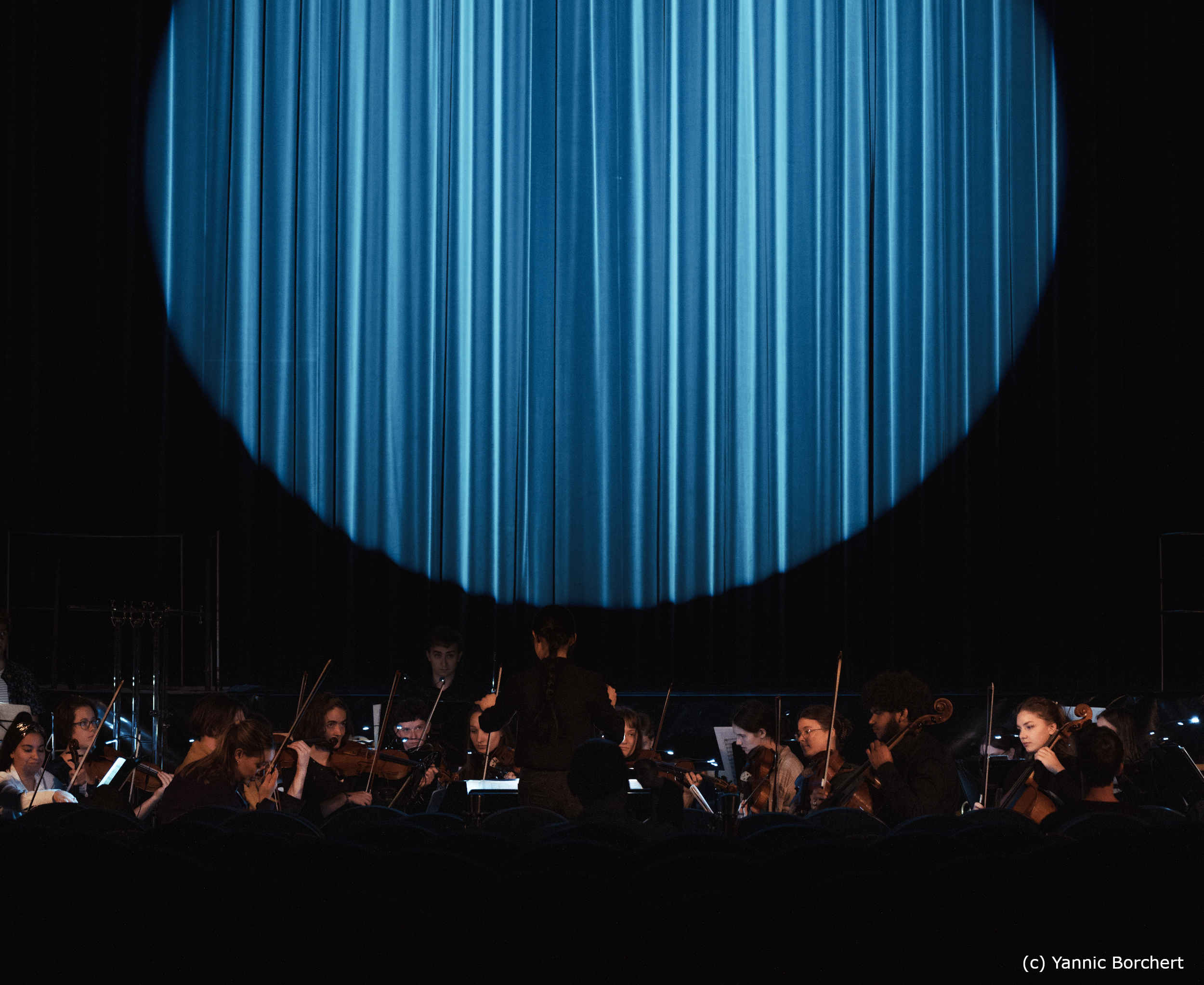 Orchester spielt im Vordergrund, im Hintergrund ist ein kreisförmiger Ausschnitt eines blauen Bühnenvorhangs