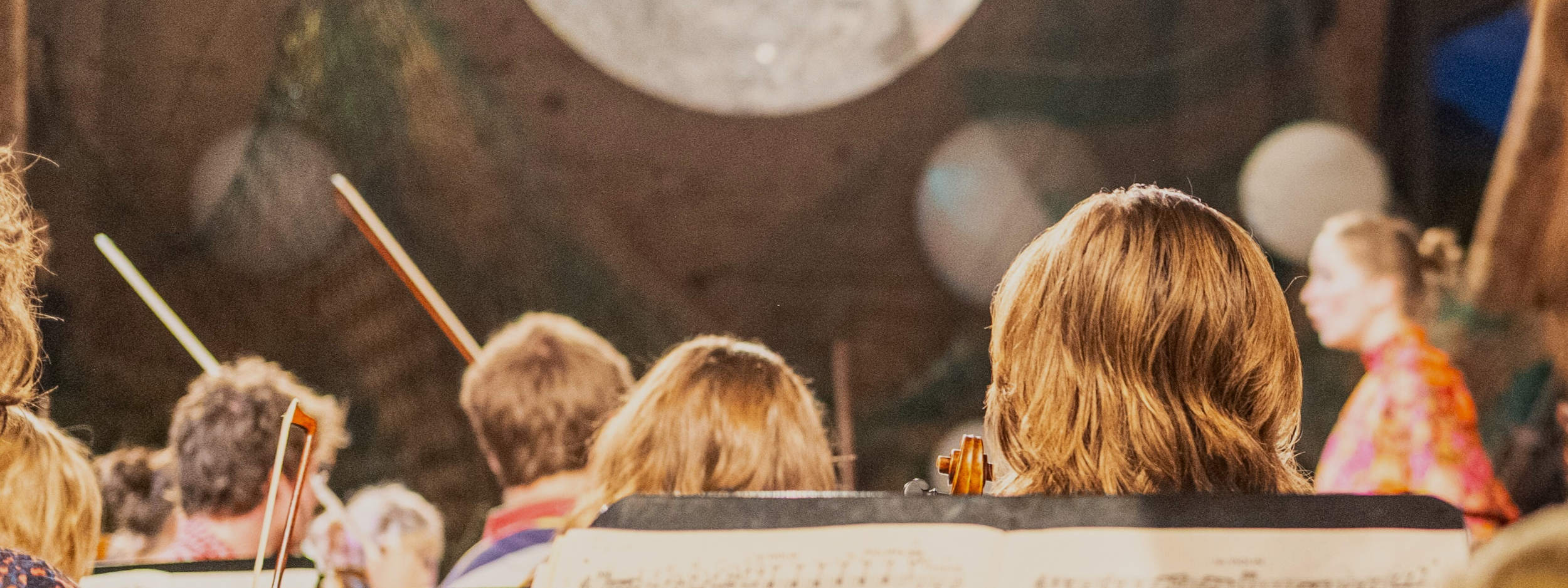 Musikerinnen und Musiker sichtbar von hinten aus dem Orchester heraus fotographiert, im Hintergrund ist ein Mond projeziert