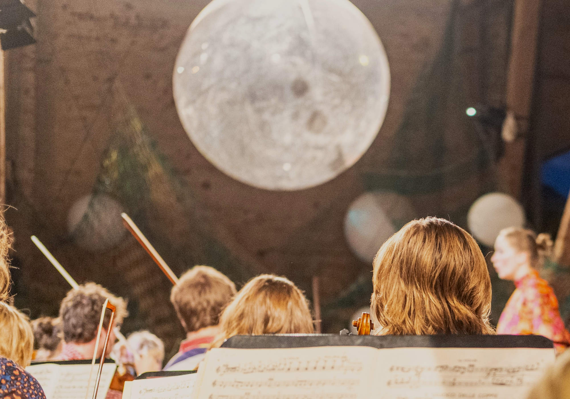 Musikerinnen und Musiker sichtbar von hinten aus dem Orchester heraus fotographiert, im Hintergrund ist ein Mond projeziert