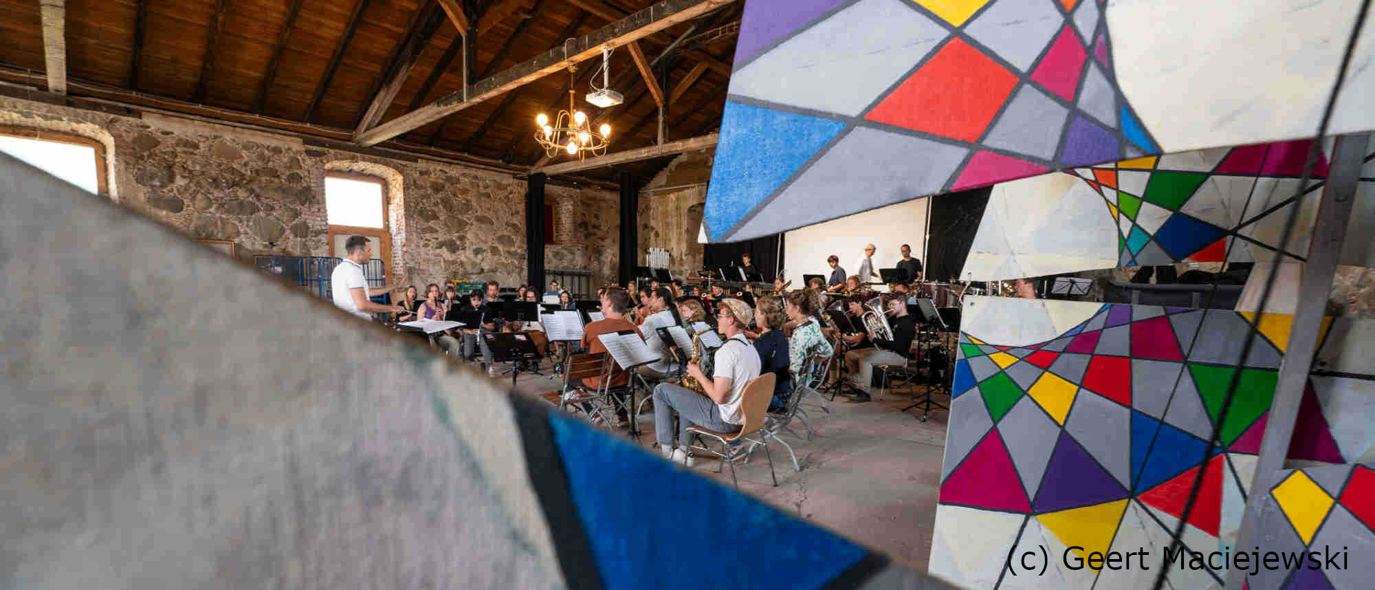 Im Vordergrund befindet sich eine Kunstinstallation, im Hintergrund sieht man ein Orchester das gerade probt.